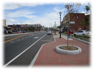 This image shows a completed construction project located in an urban area with several Complete Streets elements including: shared bike lanes, sidewalks, on-street parking, decorative lamp posts, and street trees.